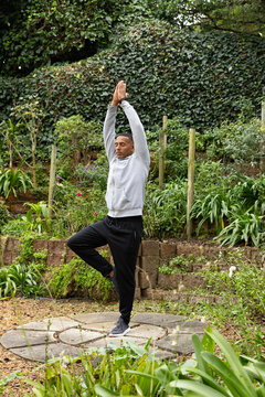 Man practicing standing yoga on circular stone platform in backyard garden wearing gray hoodie