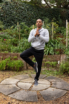 African man standing on circular stone platform in garden holding hands at chest, wearing hoodie