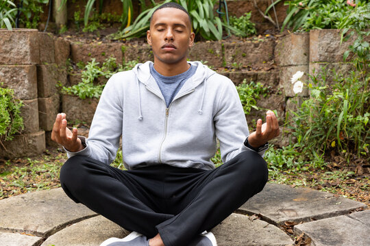 Adult African American man sitting legs crossed meditating in backyard on stone slab wearing hoodie