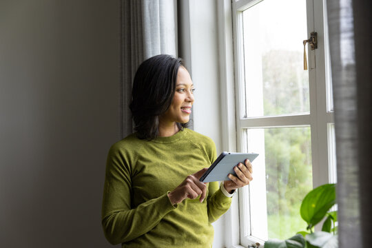 Adult African woman standing by white paned window in olive sweater using tablet, plant on sill