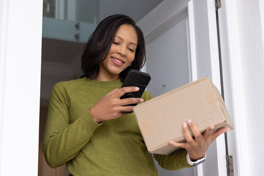 African American woman smiling, checking smartphone and holding package at door in olive sweater