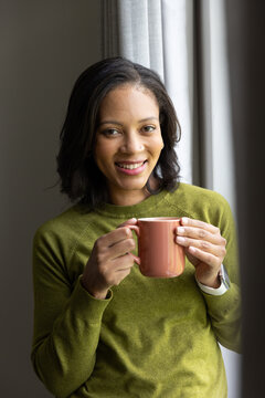 African American woman standing by tall window holding peach mug in moss green sweater, smiling