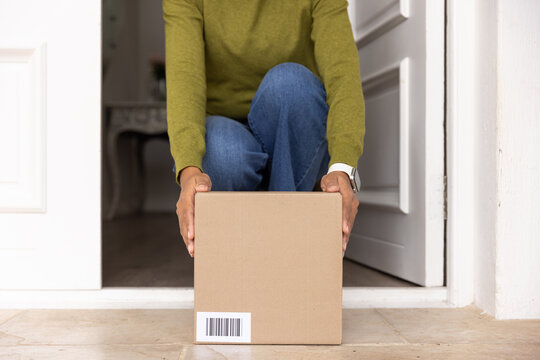 African American woman kneeling at doorway holding cardboard box with barcode, in sweater and watch