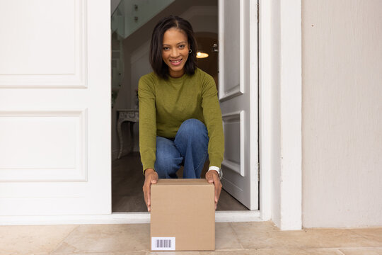 African American woman in sweater kneeling, placing sealed parcel on tiled doorstep at home