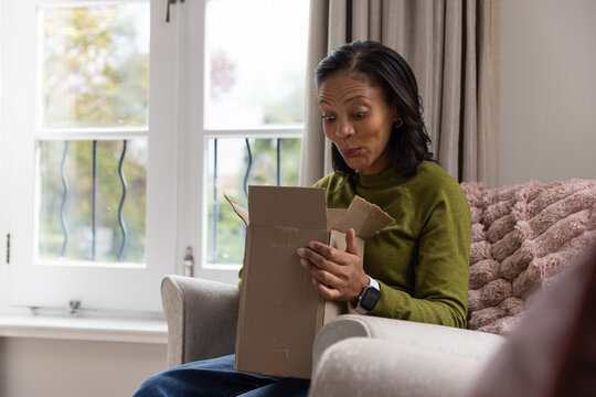 African American woman in her 40s sitting by window opening cardboard box, wearing smartwatch