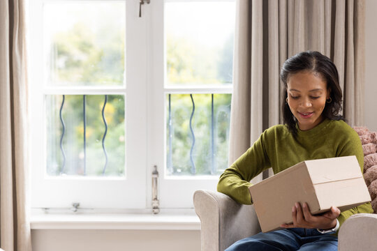 African woman wearing olive sweater jeans sitting in chair by window opening small box, copy space