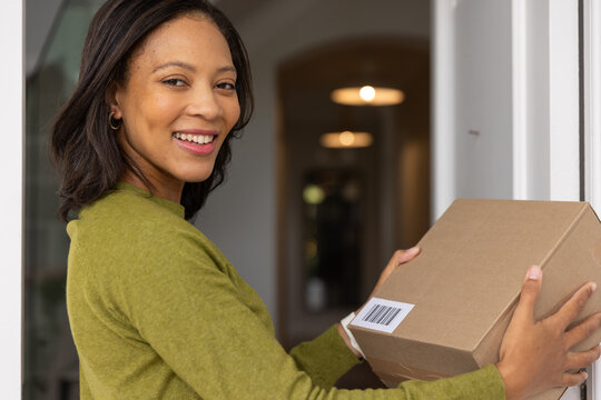 African woman holding parcel with barcode at front doorway wearing green sweater, hoop earrings