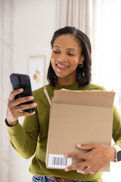 African American woman holding shipping box with barcode at chest checking phone at home