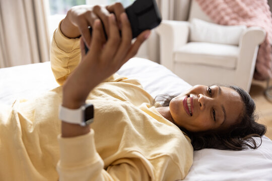 African American woman lying on bed at home in yellow sweatshirt holding smartphone and smartwatch