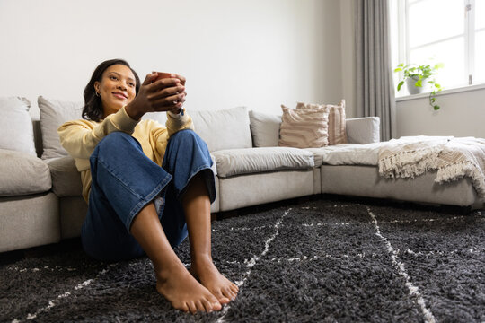 Adult African American woman sitting on rug by sofa holding mug wearing yellow top, copy space