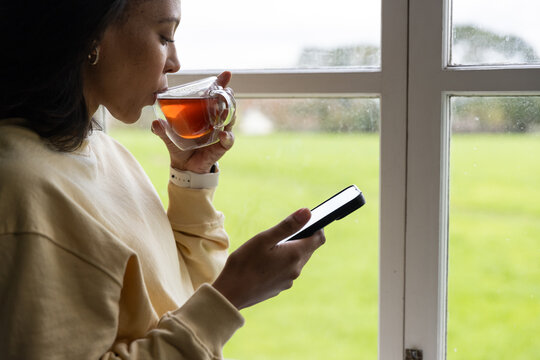 Woman standing by wet window sipping tea in glass, using phone wearing sweatshirt watch, copy space