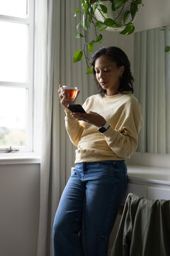 African American woman leaning at vanity by curtained window, holding glass tea and checking phone