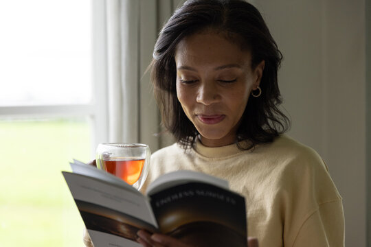 African American woman reading paperback with glass cup of amber tea in beige sweater near window