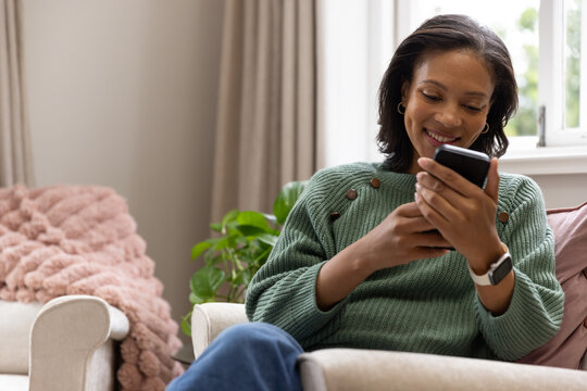 African American woman sitting on light chair at home in green sweater holding phone and smiling