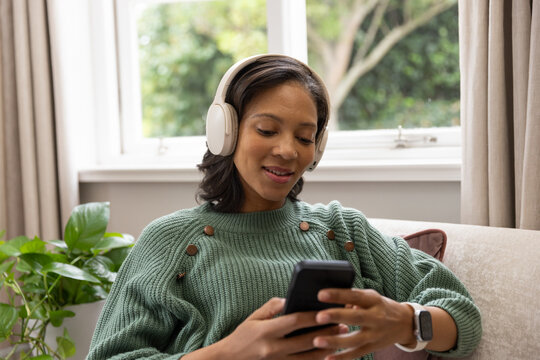 African American woman sitting on beige sofa in green sweater holding smartphone, white headphones
