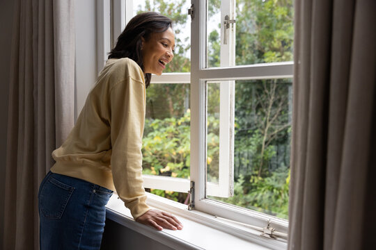 African American woman leaning on white sill by open window, wearing beige top, jeans, smiling