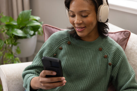 Woman sitting in cozy living room on light armchair wearing beige headphones holding smartphone