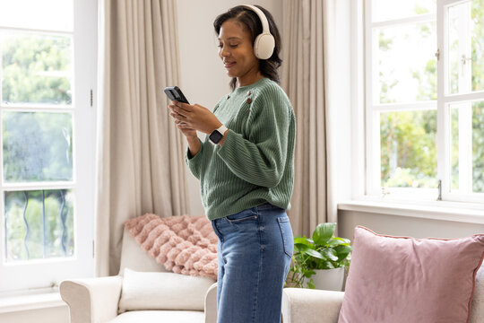 African American woman standing and using smartphone while wearing white headphones at home