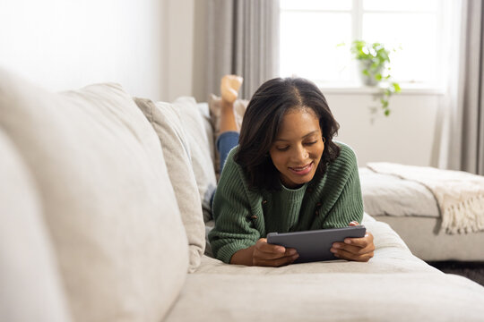 African American woman lying on beige sofa wearing green sweater holding tablet in living room