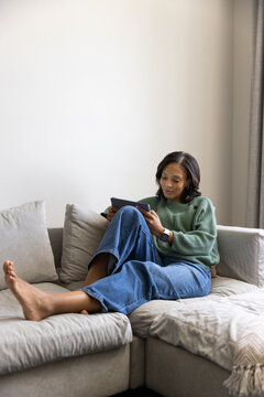 African American woman reclining on L sofa holding tablet, wearing green sweater, copy space
