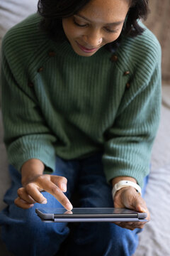 African American woman sitting at home on sofa wearing green sweater, scrolling tablet
