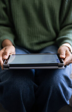 Woman holding tablet in folio case while sitting on sofa, wearing green ribbed sweater and jeans
