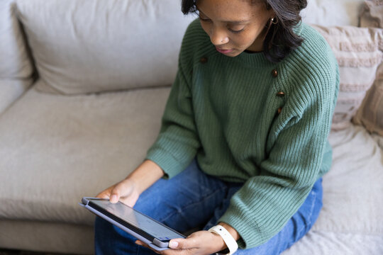 Woman sitting on light beige sofa at home holding tablet in folio case, wearing green sweater