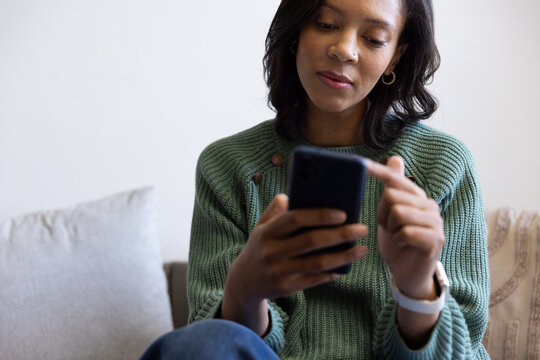 Woman holding smartphone and scrolling on sofa in living room, wearing green knit sweater