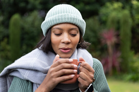 Adult African American woman standing chest-up in garden, holding terracotta mug, wearing beanie