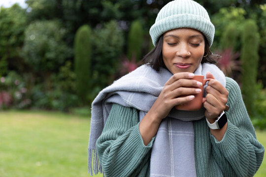 African American woman holding mug near face, inhaling in garden with beanie, scarf and smartwatch