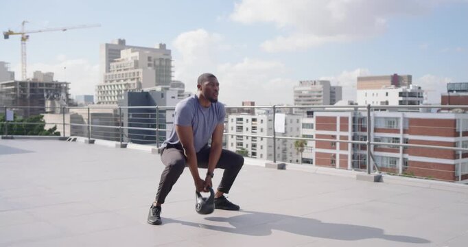Mid-adult man in light-blue tee starting wide squat, doing kettlebell swings on rooftop for fitness