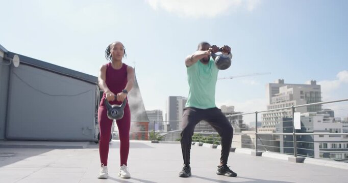 Fit pair exchanging smiles, lifting kettlebells overhead on rooftop, resting, prepping next set