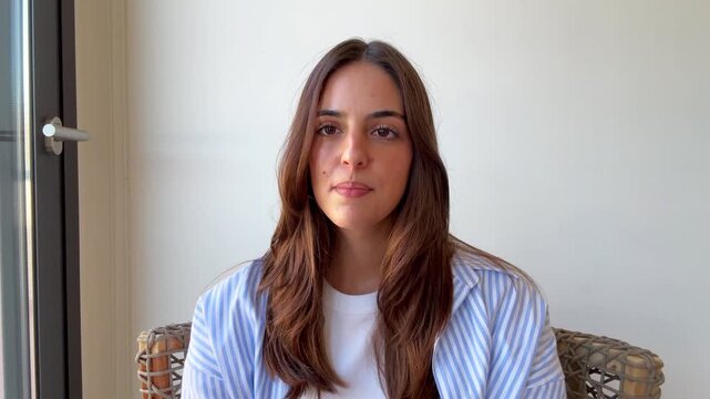 Confident young woman talking directly to the camera during a video conference call, participating in an online interview or webinar from her home office with natural window light