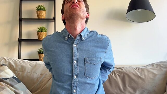 Young caucasian man sitting on the sofa at home with an expression of intense pain, suffering from a sudden backache while trying to stretch his sore muscles in the lumbar region