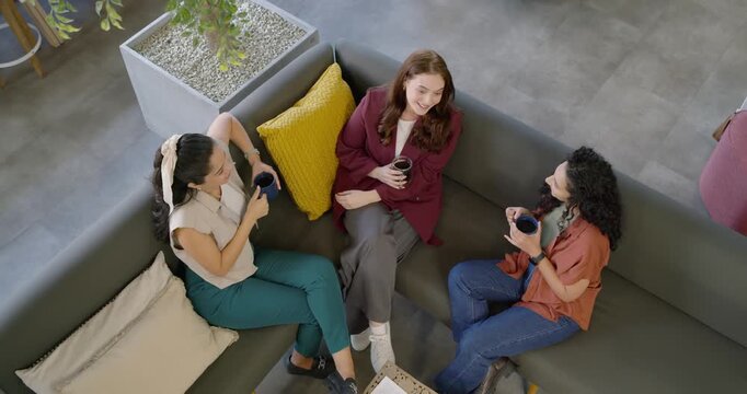 Female friends sitting on L-shaped couch in lounge, center woman starting chat, others sipping mugs