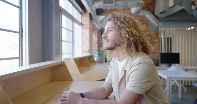 Man 20s sitting wood counter large windows wearing beige shirt taking breath calming then laughing