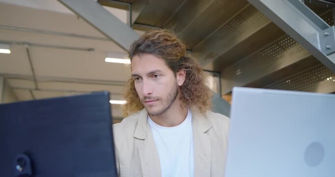 Man in his 20s working at desk in office with screens, shifting gaze, resting chin, typing
