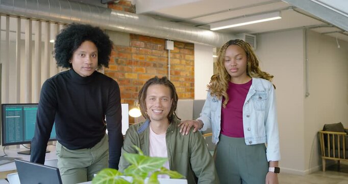 Diverse coworkers posing at desk with laptop, camera zooming, moving closer, placing hand, smiling