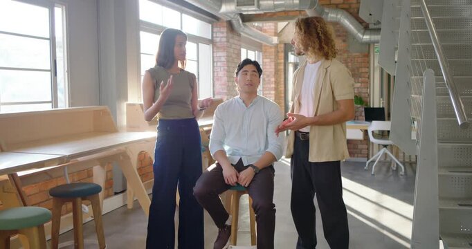 Diverse coworkers responding to idea in sunlit lounge, discussing with stool nearby, smiling