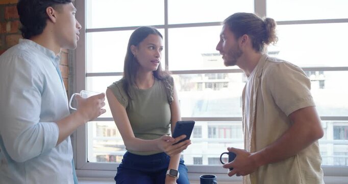 Diverse team leaning on loft ledge, woman showing phone, men holding mugs, gesturing and reacting