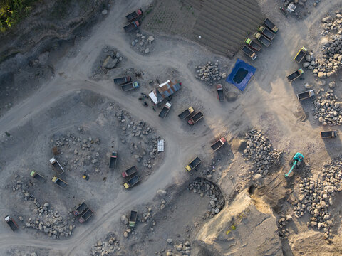 Aerial view of a sand and stone quarry with numerous dump trucks parked on dirt paths, piles of rocks, and heavy machinery in Manisrenggo, Central Java, Indonesia.