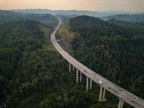 Aerial view of Jalan Tol Bawen - Salatiga highway bridge winding through lush green hills and dense forest in Sidorejo, Central Java, Indonesia.