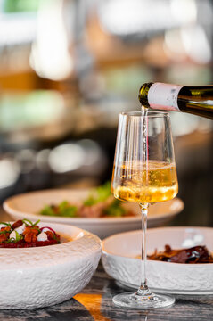 White wine pouring into a crystal glass on a restaurant table with gourmet food plates, close-up.