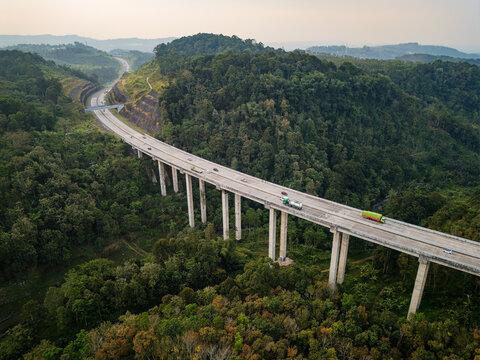 Aerial view of the Jalan Tol Bawen - Salatiga elevated highway bridge winding through lush green forests and hilly terrain in Sidorejo, Central Java, Indonesia.