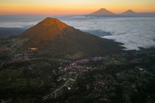 Aerial view of Mount Andong at golden hour, rising above clouds with terraced fields and villages in the valley, Ngablak, Central Java, Indonesia.