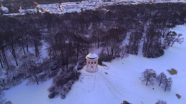 Cinematic aerial view of the historic Monopteros rotunda in Englischer Garten, Bavaria, featuring a snow-covered sledding slope and public park landscape in crisp morning light of a winter dawn.