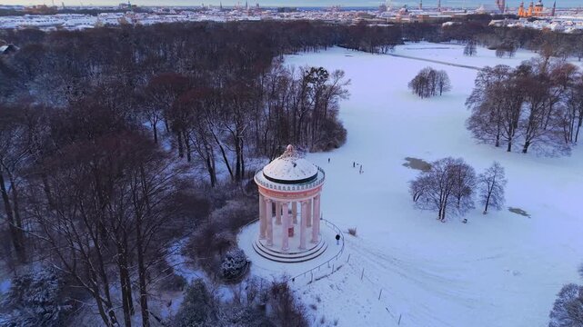B-roll drone footage capturing the quiet winter morning at Munich's famous Monopteros landmark, with golden sunrise light illuminating the snow-covered public park meadows and sleeping trees.