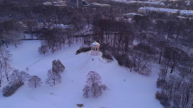 Beautiful winter drone perspective of the frozen Monopteros pavilion on an artificial hill in Munich, Germany, capturing the golden dawn light on snowy trees and a famous historical viewpoint.