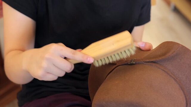 A wooden brush removes dust from a brown felt hat on a person's lap, showing simple indoor care for a personal accessory.