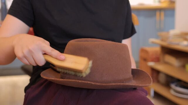 A person brushes a brown felt hat indoors, removing dust from the brim and crown during a calm wardrobe care routine.